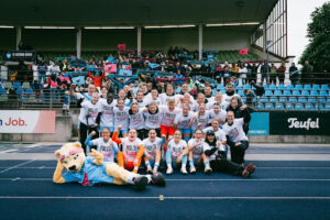 Das Team des FC Viktoria Berlin auf der blauen Tartanbahn des Stadions vor der Tribüne mit Fans. Die Spielerinnen & der Staff tragen alle weiße T-Shirts mit der Aufschrift: Hallo 2. Liga!