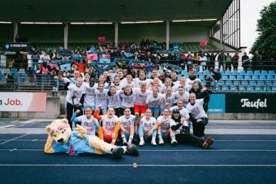 Das Team des FC Viktoria Berlin auf der blauen Tartanbahn des Stadions vor der Tribüne mit Fans. Die Spielerinnen & der Staff tragen alle weiße T-Shirts mit der Aufschrift: Hallo 2. Liga!