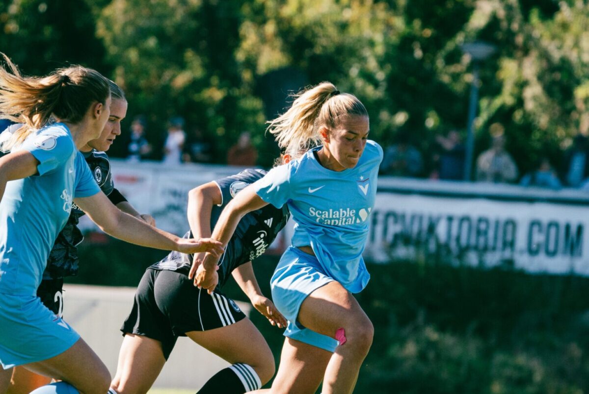 Nina Ehegötz im himmelblauen Viktoria-Berlin Trikot, die auf den Boden nach dem Ball schaut. Hinter eine kaum zu erkennende Frankfurterin im grauen Trikot, links am Bildrand eine Viktoria-Mitspielerin.