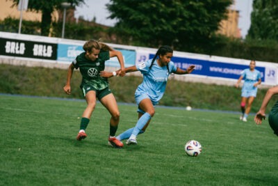 Die FC-Viktoria-Berlin-Spielerin Maya Hahn mit zwei langen geflochtenen dunklen Zöpfen und himmelblauem Trikot im Zweikampf mit einer Wolfsburgerin im dunkelgrün/hellgrünen Trikot.